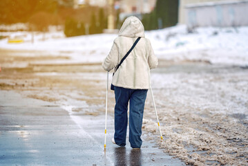 Senior woman with walking canes in hands walks along snowy sidewalk in winter city street. Old...
