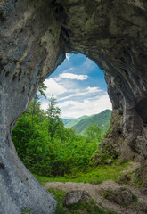 Viewpoint above a deciduous forest and mountains through a cave, eroded in a calcareous cliff on a mountain side. The tunnel has its stone walls covered with moss and lichen. Natural phenomenon.