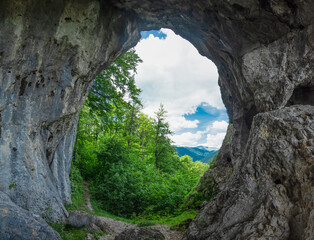 Viewpoint above a deciduous forest and mountains through a grotto, eroded in a calcareous cliff on a mountain side. The tunnel has its stone walls covered with moss and lichen. Natural phenomenon.