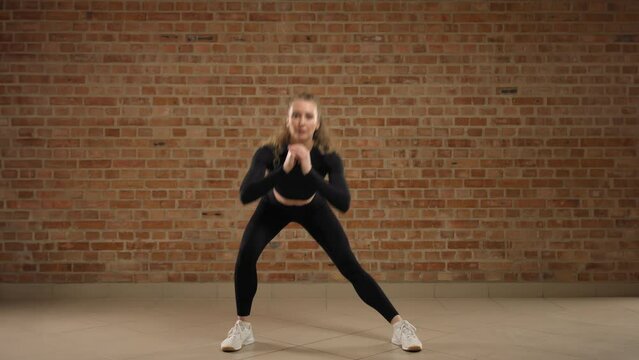 A fitness trainer performs an exercise, side squats. She clearly demonstrates the move on camera, doing so professionally. The trainer is dressed in black athletic wear. RED V-RAPTOR 8K RAW.