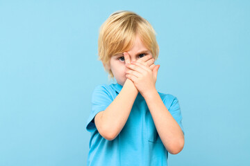 Emotional young boy covering face with hands isolated over pastel blue background. Mental wellbeing and safety in childhood age.