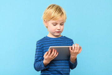 Young child holding digital tablet. Boy watching videos on his computer, isolated over pastel blue...