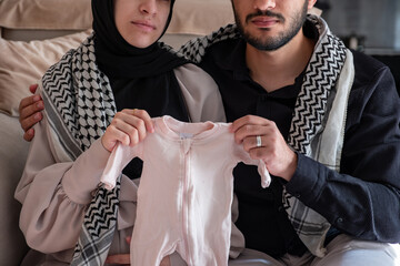 Husband and his wife holding newborn clothes for there dead child  while wearing palestinian keffiyeh in living room