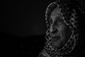 portrait of an old lady in dark background wearing white palestinian keffiyeh with smile on her...