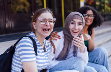 Multiracial female students laughing outfoors. Happy friends with backpacks have fun and relax.