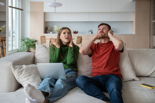 Angry annoyed couple sitting on sofa, suffering from noisy neighbors, closing ears. Wife and husband spending weekend at home, having no opportunity to rest, work because of irritating sound, knock