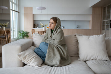 Sad tired woman sits on couch at home wrapped in plaid in living room. Blue mood of depressed girl...