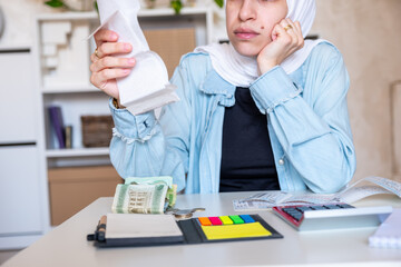 Young Female wearing hijab sitting in the living room checking receipts and using calculator to...