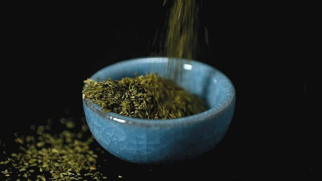 Hand sprinkling pouring seasoning oregano basil in blue bowl on black background. slow motion