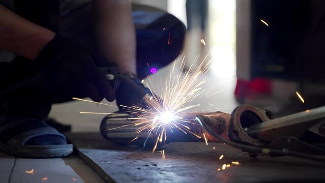 Auto mechanic welds car exhaust flex pipe, catalyst in garage. Welding sparks fly from repair work on vehicle underside. Professional service for automotive exhaust system maintenance.