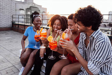 Company of four cheerful friends are enjoying tasty cocktails. Roof party. Interracial man and african women drink aperol with orange slices.