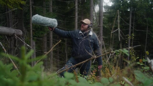 Caucasian man wearing headphones records sounds of nature for movie in coniferous wood using professional audio equipment. Sound designer works with furry windshield microphone outdoors. Filmmaking.