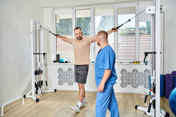 young physiotherapist assisting male patient working out on exercise machine in kinesio center - obrazy, fototapety, plakaty