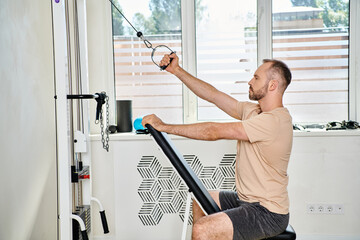 side view of handsome man in sportswear working out on training machine in kinesiology center - obrazy, fototapety, plakaty