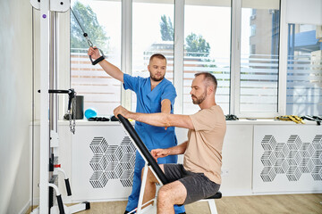 male patient in sportswear training on exercise machine near young doctor in kinesio center