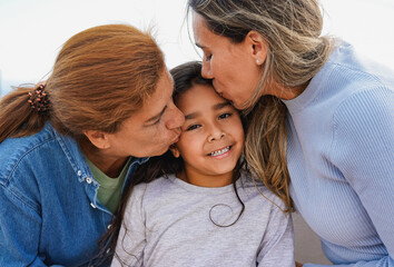 Latin multi generational family having tender moment outdoor - Female child smiling on camera while mother and grandmother kissing her face - obrazy, fototapety, plakaty