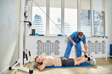 physician in blue uniform assisting man working out on exercise machine in gym of kinesio center