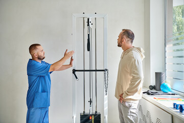 rehabilitologist in blue uniform showing exercise equipment to his patient in kinesio center