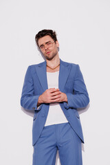Confident and stylish brunette man in blue suit and eyeglasses looking at camera on grey background