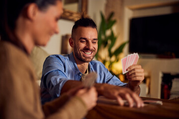 Focus on the smiling man playing cards with his girlfriend, at home.