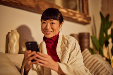 Portrait of a smiling Asian woman, using a mobile phone, looking at the camera while resting on the couch. - obrazy, fototapety, plakaty