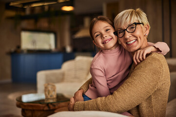 Portrait of a grandchild and her grandma smiling for the camera while sitting on the couch in the living room.