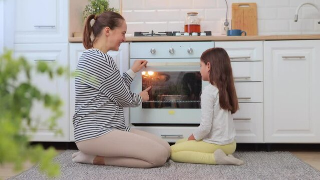 Brown haired mother and daughter sitting on floor near oven waiting for tasty dessert together talking and laughing enjoying family moments on weekend