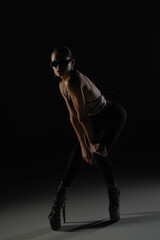 Fototapeta na wymiar Young woman in black bodice and tights poses on black backlit studio background. Female dancer demonstrates elements of dance on high heels. Dance promo video concept.