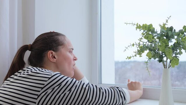 Sad bored woman with brown hair sitting near window touching glass yawning being sleepy tired and lonely female wearing striped shirt spending boring time at home