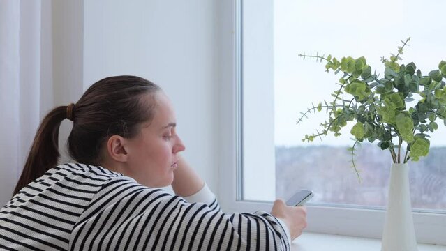 Bored sad woman wearing striped shier sitting near window with her smartphone using mobile app checking emails and social networks trying to entertain herself with internet