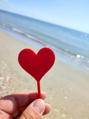 Fototapeta premium Person holding in fingers hand stick in shape red heart on background sea and sea waves, blue sky in seashore on sunny summer day close-up. Concept love romance amour St Valentines Day