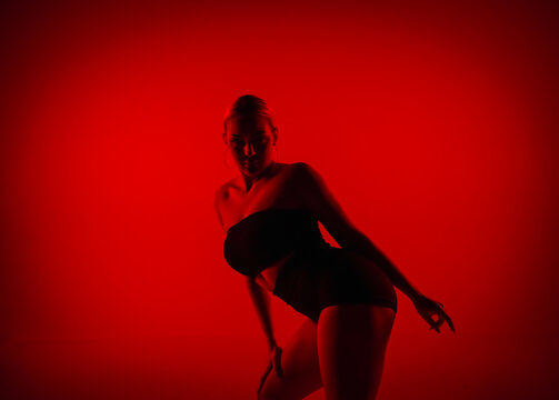 Silhouette female dancer in black shorts and top dancing on high heels. Young woman poses gracefully and shows off slender flexible body in dark studio with red light.
