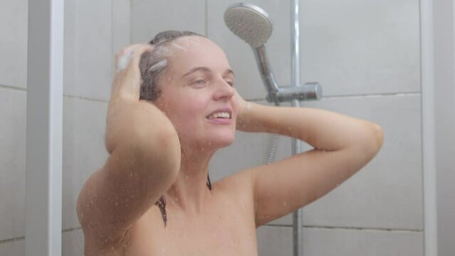 Caucasian young adult naked woman washing her hair in shower with shampoo foam enjoying refreshing and cleansing routine in the bathroom cabin at home