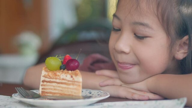 Closed up face of hungry asian kid girl, faces with innocent curiosity, gather around a table with yummy, beautiful desserts. Delicious cakes, eyes wide with anticipation, leans closer for a taste. 