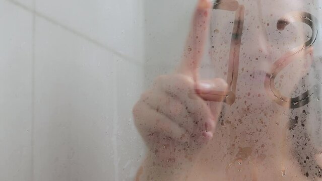 Attractive young woman writing ward sex on weeping glass shower door enjoying rest in douche washing her body standing behind steam blurred glass with water drops