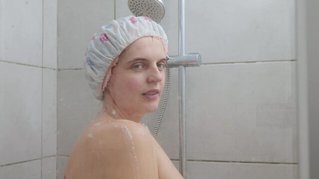 Young adult beautiful naked woman taking shower in bathroom at home wearing shower cap doing morning hygiene routine taking care of her body cleanliness