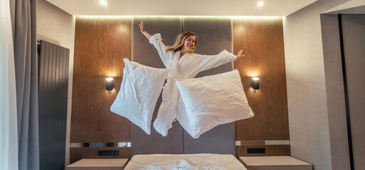 Woman having fun and jumping with bathrobe on bed in hotel room