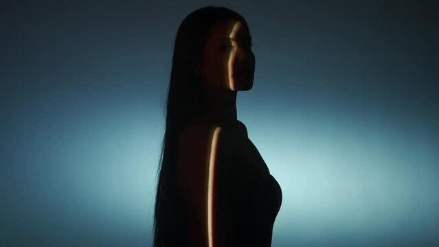 Portrait of female model in studio. Silhouette appealing woman posing on white low light background, turns at camera with selective ray of light.