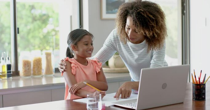 Happy biracial mother and daughter using laptop for schoolwork in kitchen, slow motion
