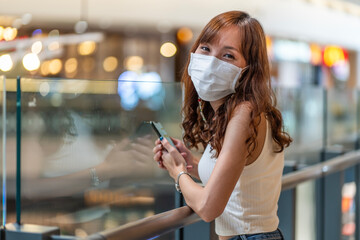 Asian woman, a customer, comes to shop in the mall wearing a face mask and holding a smartphone in...