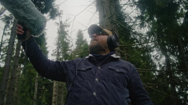 Male traveler wearing headphones records sounds of nature for movie in coniferous wood using professional audio equipment. Sound technician works with furry windjammer microphone outdoors. Filmmaking.