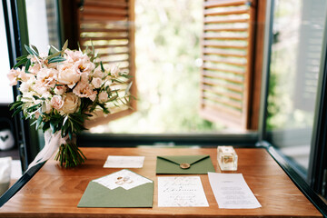 Bouquet of flowers stands near invitations and wedding rings on a wooden table by the window