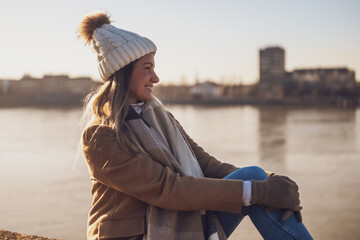 Beautiful woman in warm clothing enjoys resting by the river on a sunny winter day. Toned image.
