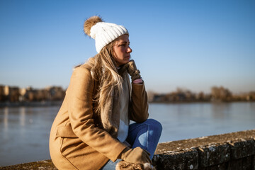 Sad woman in warm clothing sitting by the river on a sunny winter day.