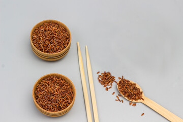 Red brown rice in wooden bowls and coconut shells. Wooden spoon with rice and chopsticks on gray