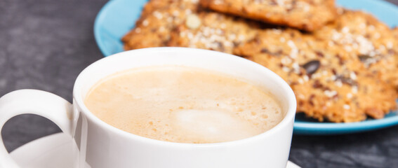 Cup of coffee with milk and fresh baked oatmeal cookies with honey and healthy seeds on blue plate. Delicious crunchy dessert