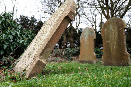 Shallow focus of a stone grave seen heavily leaning over due to ground subsidence in an English rural cemetery. Soon the gravestone will fall flat on the grass.