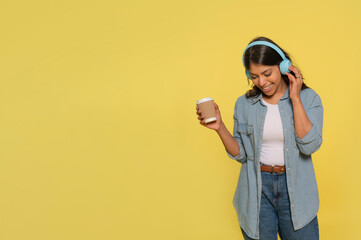 Happy young Indian woman carried away with music dances carefree with arms raised  wears stereo headphones on her ears dressed in jeans and a denim shirt isolated on  yellow background