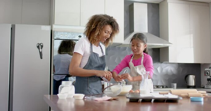 Happy biracial mother and daughter making cake mix, baking in kitchen, slow motion