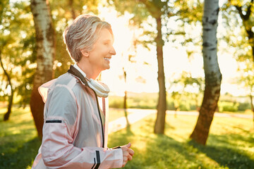 Exercises on fresh air. Side view of cheerful active woman with short grey hair running alone at green city park with wireless modern headphones on neck. Beautiful summer sunrise on background.